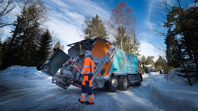 Bilde av en søppelbil på vinterføre, hvor en arbeider står i orange arbeidsklær og tømmer en avfallsdunk inn i søppelbilen.